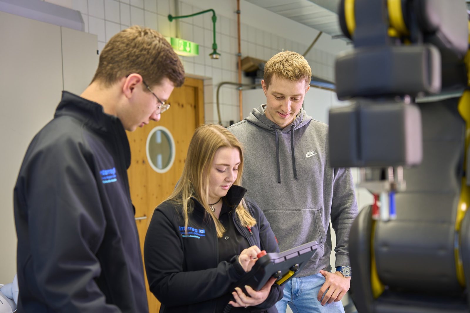 Three young people at a robot cell; a woman operates a tablet, a robot arm in the foreground.