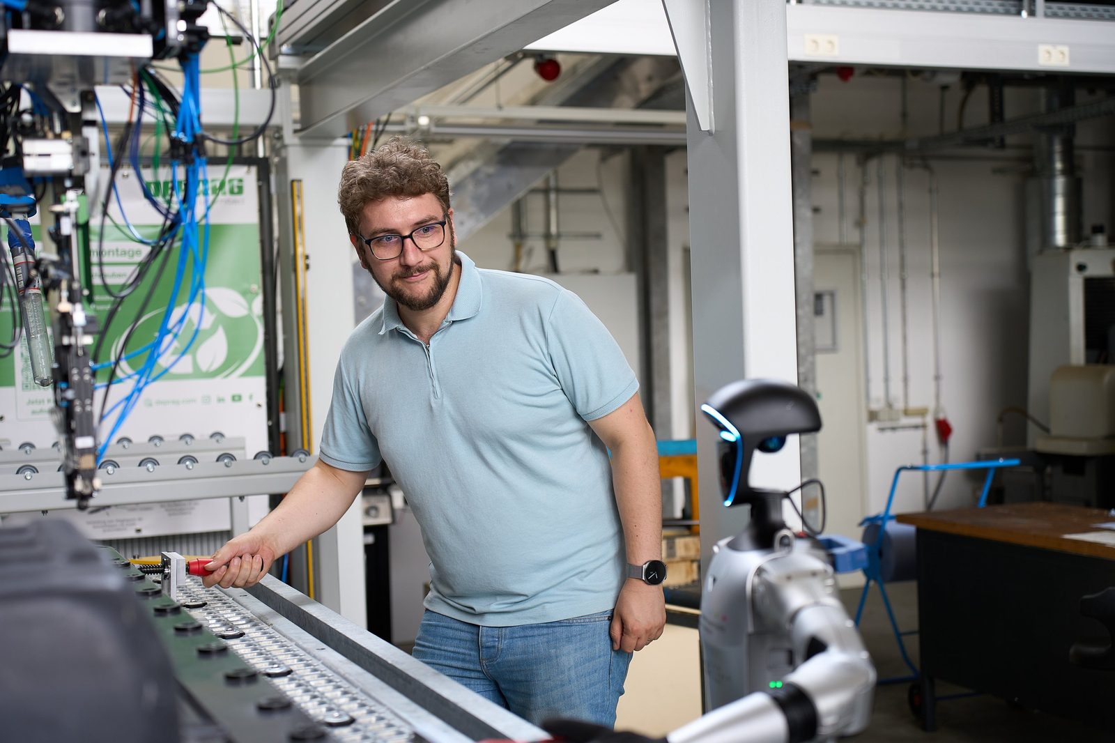A man at the control panel of an automated assembly line; a humanoid robot stands in the background.