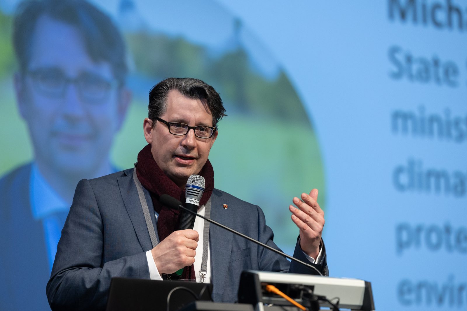A man with glasses and a scarf speaks into a microphone in front of a projection.