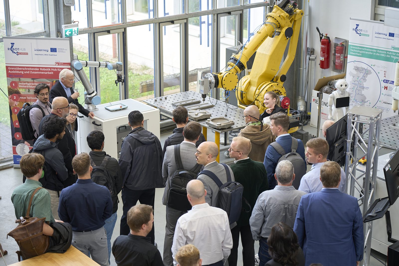 A large group of participants around a yellow industrial robot in the lab hall.