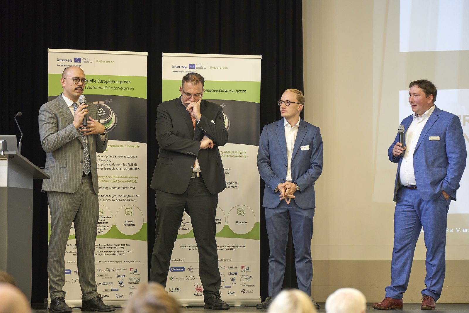 Four men on stage in front of PAE-e-green roll-ups, one speaking into a microphone.