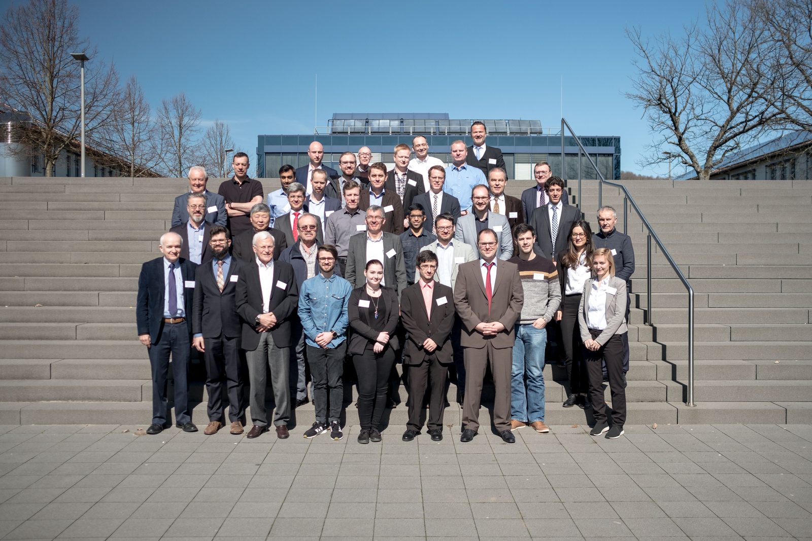 Group photo of about fifty participants on the outdoor steps of the campus in spring sunshine.
