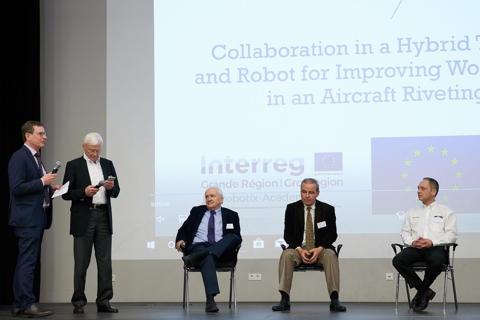 Panel on stage: three men seated, two standing with microphones.